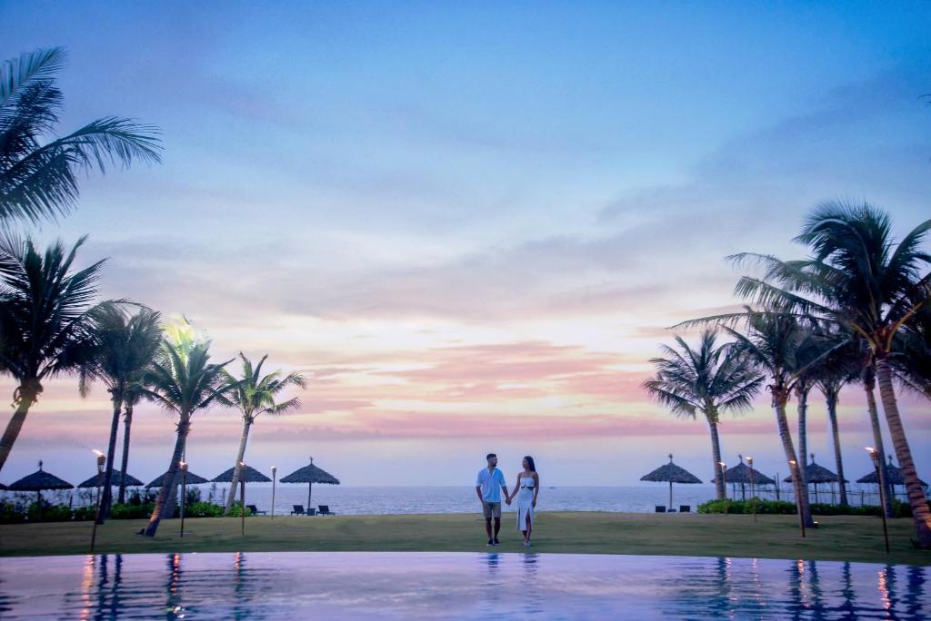 会安Wyndham Hoi An Royal Beachfront Resort & Villas的日落时分一对夫妇站在海滩前