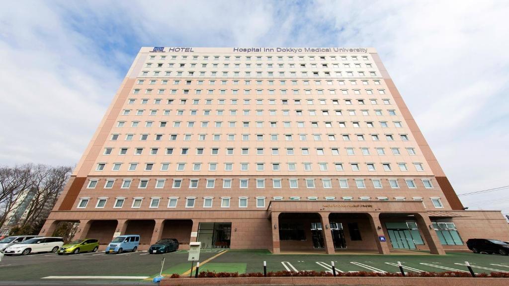 MibuToyoko Inn HOSPITAL INN Dokkyo Medical University的一座高大的建筑,前面有汽车停放