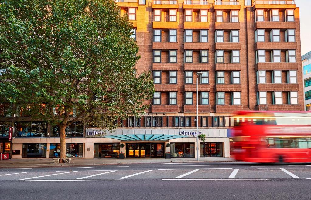 伦敦希尔顿伦敦奥林匹亚酒店的一辆红色的公共汽车驶向大楼前的街道
