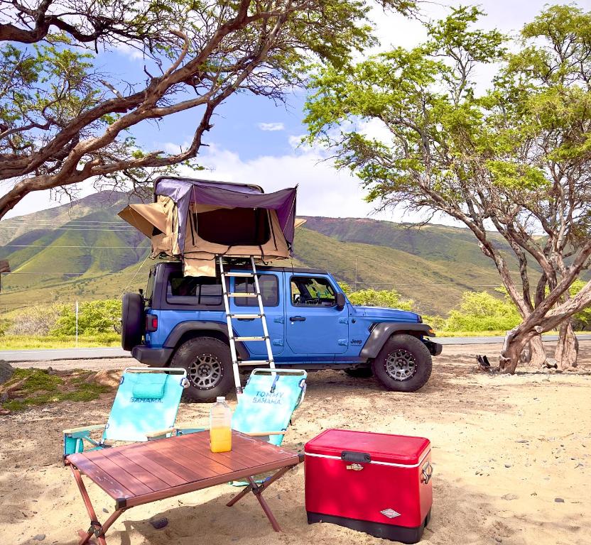 帕依亚Embark on a journey through Maui with Aloha Glamp's jeep and rooftop tent allows you to discover diverse campgrounds, unveiling the island's beauty from unique perspectives each day的一辆蓝色的卡车,带帐篷和两把椅子及一张桌子