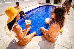 坎昆Nomads Party Hostel的两个女人坐在游泳池旁边,喝着饮料