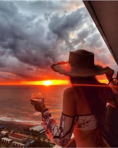 巴统PANORAMA Orbi Beach Resort Center Suite的戴着帽子的女人,一边欣赏日落,一边喝着葡萄酒