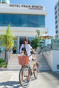 马塞约Hotel Praia Bonita Jangadeiros Pajuçara的一名妇女在建筑物前骑着自行车