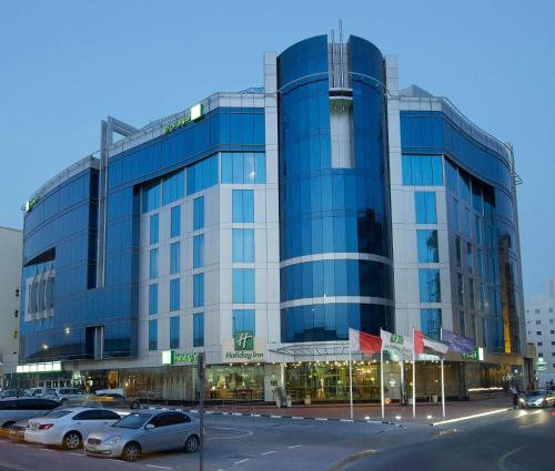 迪拜迪拜阿尔巴沙假日酒店的一座大型玻璃建筑,前面有汽车停放