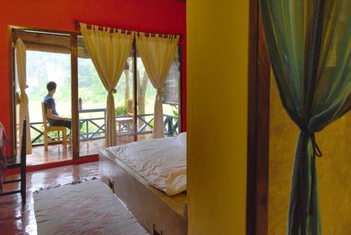 Nongkhiaw曼陀罗欧度假酒店的一间卧室配有一张床,而另一位坐在阳台上椅子上的人