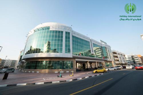 迪拜Holiday International Hotel Embassy District的一座大型玻璃建筑,前面有汽车停放