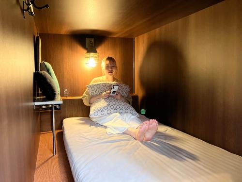 横滨HARE-TABI SAUNA&INN Yokohama的坐在床上看着手机的女人