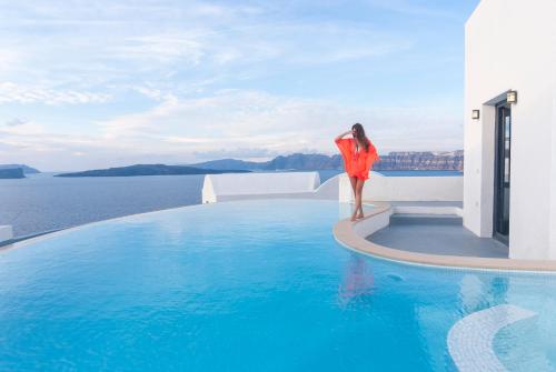 阿克罗蒂里Grand Ambassador Santorini Hotel的站在游泳池边缘的身着橙色衣服的女人